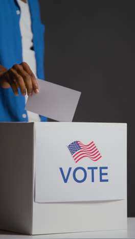 Vertical-Video-Close-Up-Of-Voters-Casting-Votes-Into-American-Election-Ballot-Box-With-USA-Flag-Against-Black-Background-Shot-In-Slow-Motion